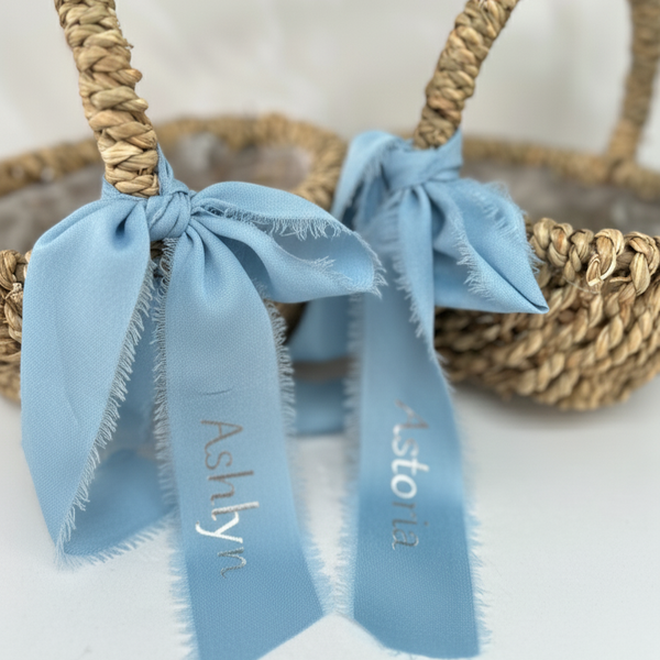 Woven flower girl basket with blue ribbons tied around it, featuring names 'Ashlyn' and 'Astor'.
