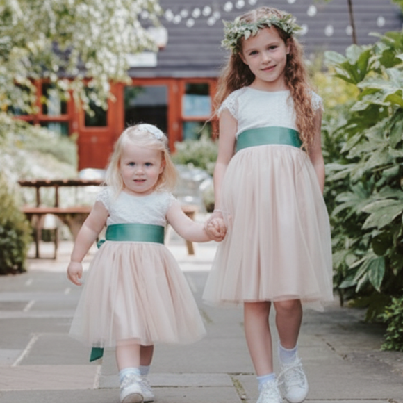 Two young girls in matching champagne dresses with green sashes walking hand-in-hand in a garden.