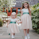 Two young girls in matching champagne dresses with green sashes walking hand-in-hand in a garden.