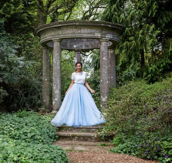 Woman in a white lace top and light blue tulle skirt standing in front of a stone gazebo in a garden.
