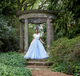 Woman in a white lace top and light blue tulle skirt standing in front of a stone gazebo in a garden.