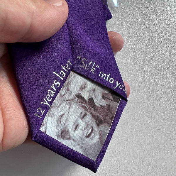 A hand holding a tie with a black-and-white portrait photo insert and silver text reading '12 years later "Silk" into you'.